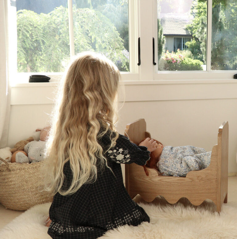 Girl playing with a handcrafted wooden doll bed and toys by a window, a basket of stuffed animals nearby