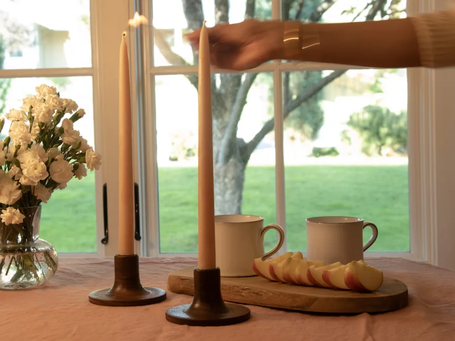 Lit taper candles in walnut holders on a table with flowers and sliced apples